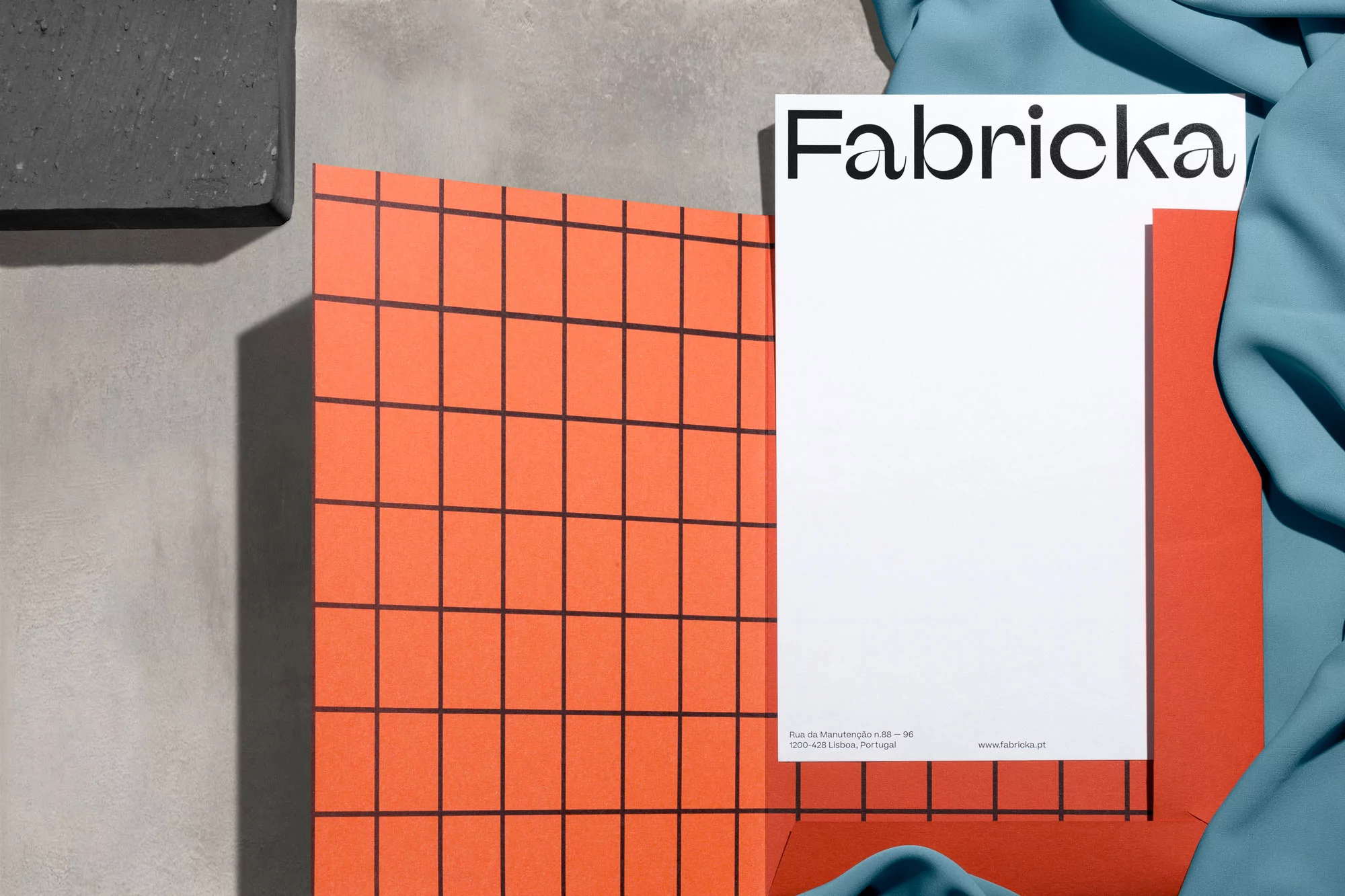 Close-up studio shot of a Fabricka flyer with an orange grid, set against a draped teal fabric and concrete surface for a graphic design layout.