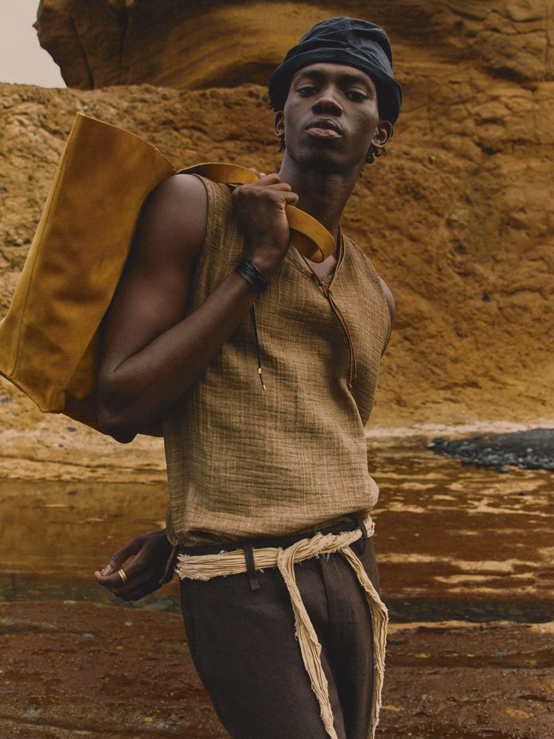 An African American male model poses outdoors in a sleeveless top and dark pants, carrying a yellow bag over his shoulder.