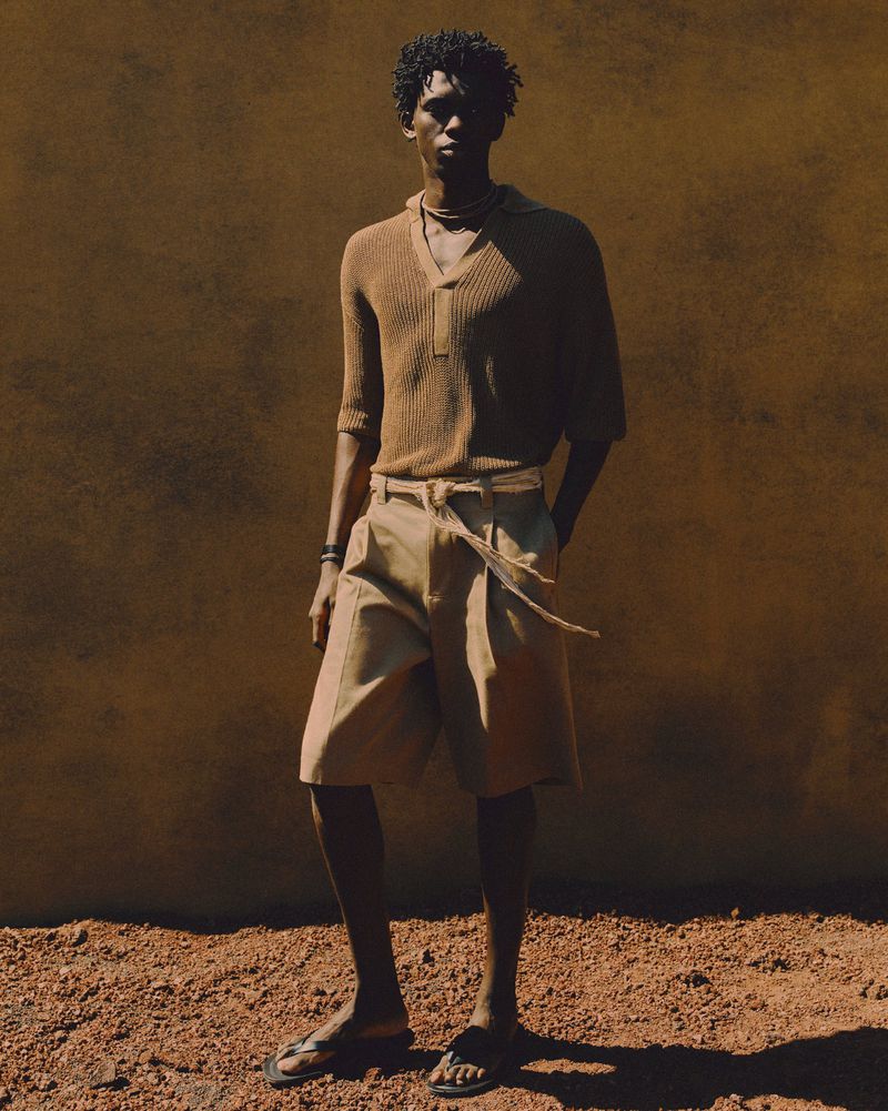 An adult male model with an afro poses in a brown knit polo, light-colored shorts, and sandals against a textured brown background in a studio shot.