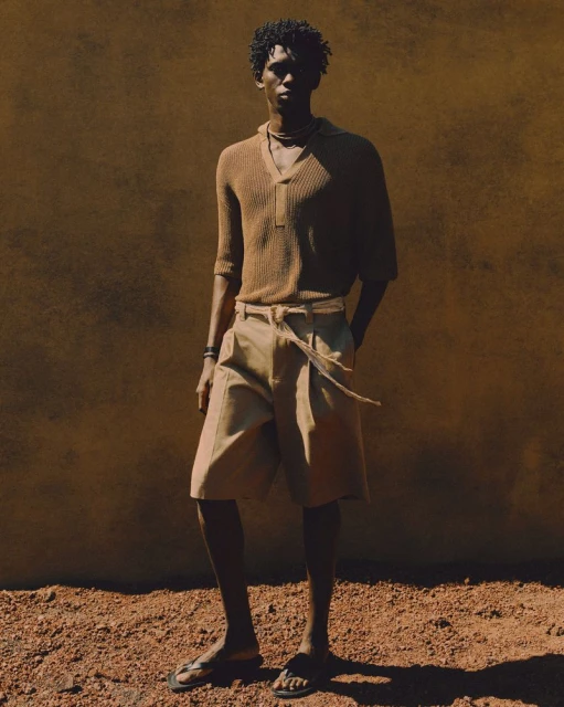 An adult male model with an afro poses in a brown knit polo, light-colored shorts, and sandals against a textured brown background in a studio shot.