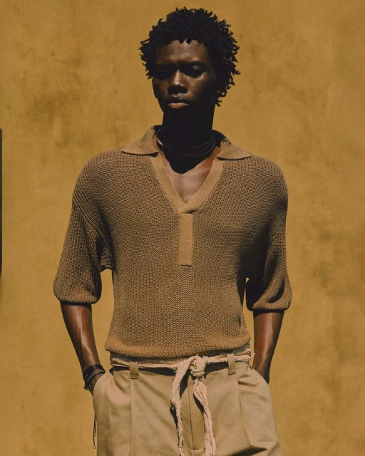 Portrait of a dark-skinned adult male model in a knit shirt and light pants against a neutral wall in a fashion or editorial style.