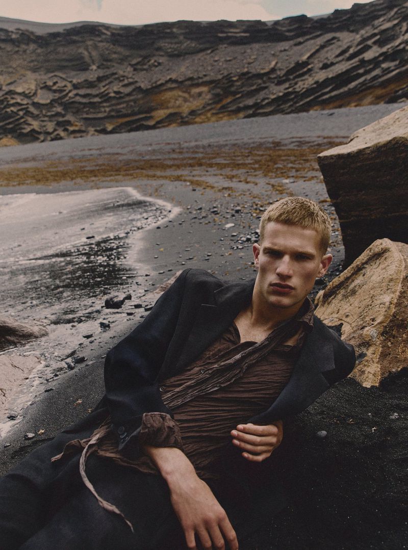 Adult male model in dark clothing poses on a black sand beach with a dark coat against a backdrop of weathered cliffs and ocean.