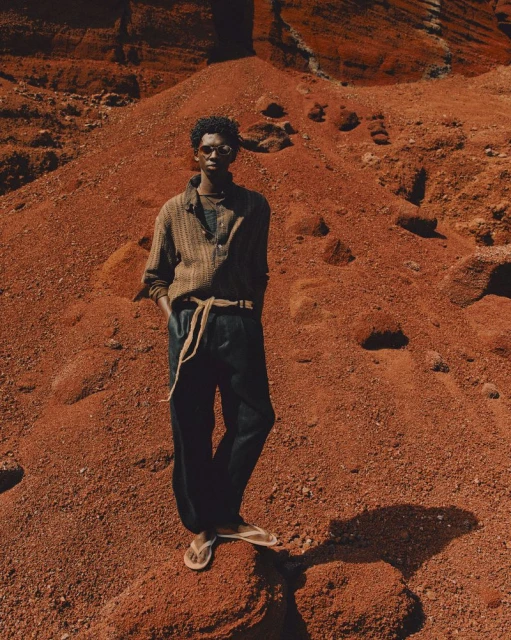 Man in casual wear stands on red desert rock formation, wearing sunglasses, button-down shirt, dark trousers, sandals, earth-tone color palette.