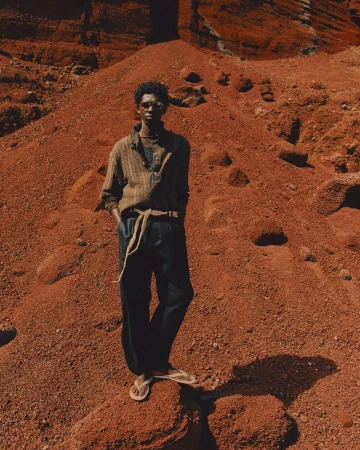 Man in casual wear stands on red desert rock formation, wearing sunglasses, button-down shirt, dark trousers, sandals, earth-tone color palette.