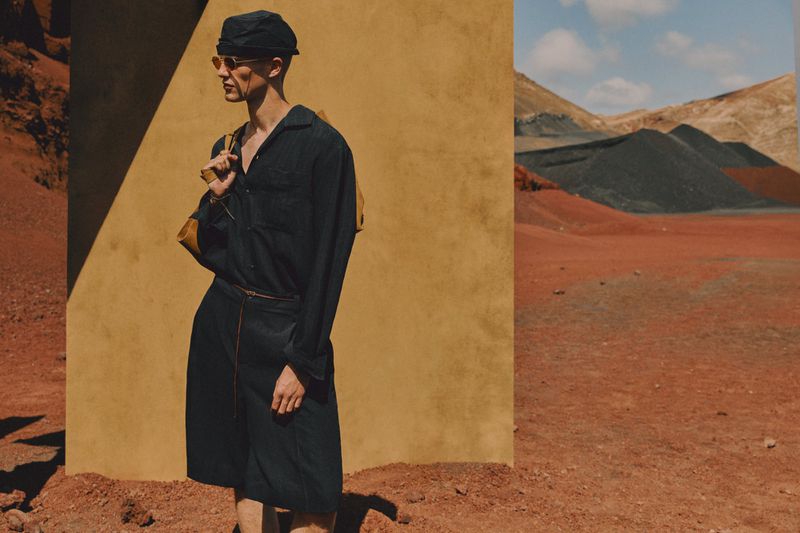 A stylish adult male stands in a desert landscape wearing a dark shirt and shorts with sunglasses and a hat, under bright sunlight.