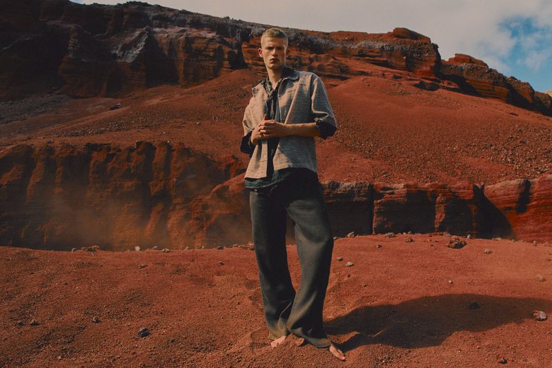 Man poses in a desert landscape wearing an open collared shirt and dark pants, under a partly cloudy sky near red rock formations.