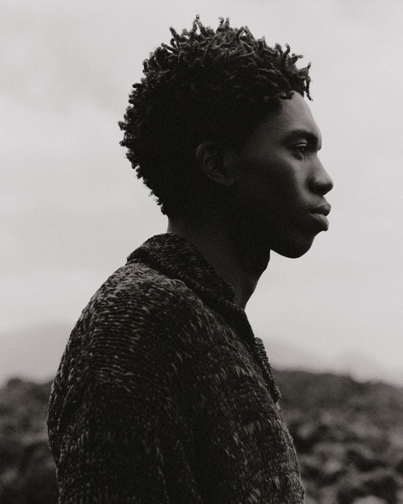 Portrait of an African American male outdoors in a tweed jacket. Side view of the man's face and upper body against a grey sky.