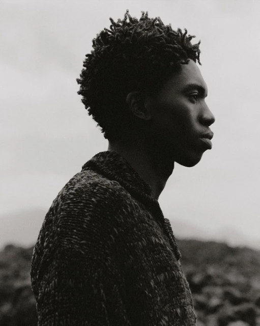 Portrait of an African American male outdoors in a tweed jacket. Side view of the man's face and upper body against a grey sky.