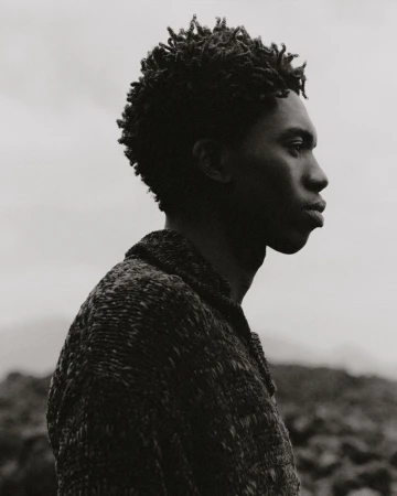 Portrait of an African American male outdoors in a tweed jacket. Side view of the man's face and upper body against a grey sky.