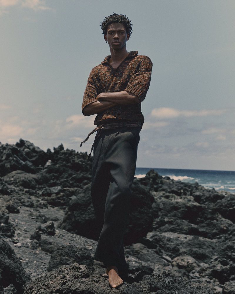 Portrait of a black male fashion model standing barefoot on a rocky beach in a knit sweater and dark pants against a cloudy sky and ocean.