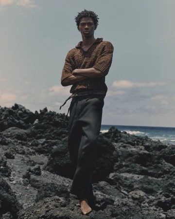 Portrait of a black male fashion model standing barefoot on a rocky beach in a knit sweater and dark pants against a cloudy sky and ocean.