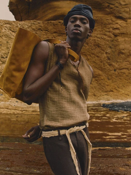 Model wearing a textured brown sleeveless top and dark cap holding a large yellow shoulder bag against a rugged rock formation.