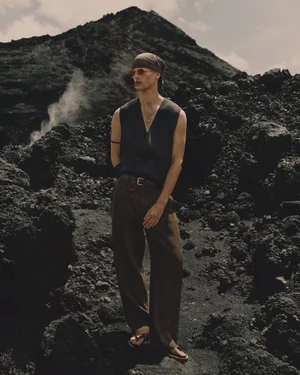 Male model in a dark vest and brown trousers standing outdoors on a dark volcanic mountain terrain.
