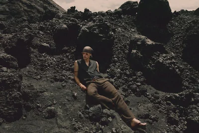 Model wearing dark sleeveless vest, trousers, and headscarf reclining on a rugged dark terrain slope under overcast sky