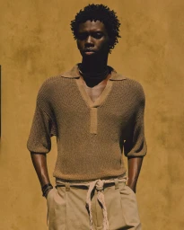 Black man wearing a brown knitted polo shirt and beige trousers against a textured golden yellow background