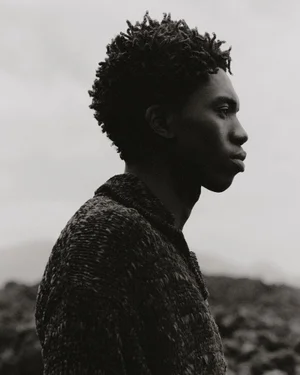 Profile portrait of a black man wearing a textured knit sweater against an overcast sky background.