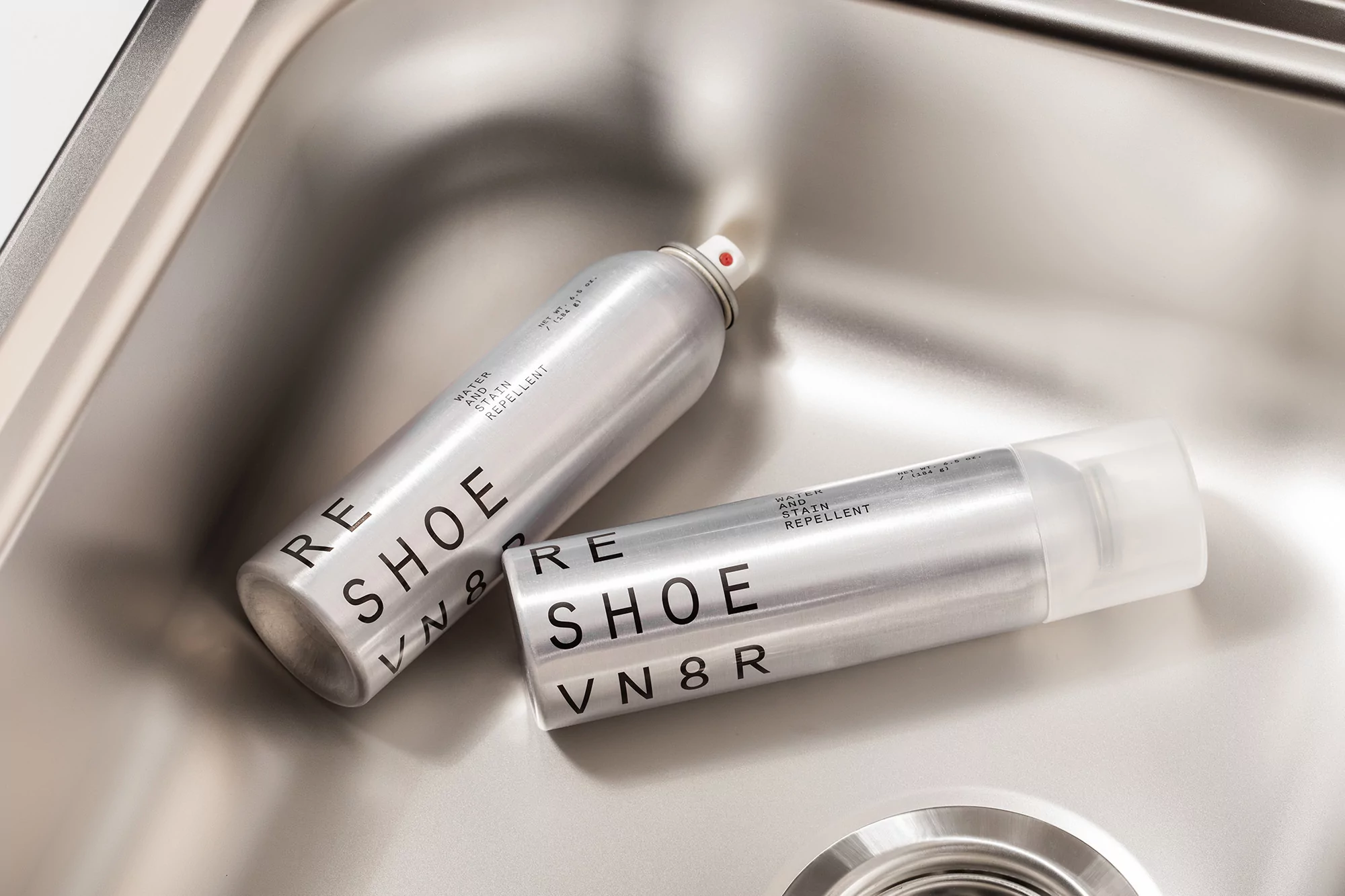 Two cans of RE SHOE VN8R water and stain repellent sit in a stainless steel sink, reflecting overhead light and highlighting the product labels.