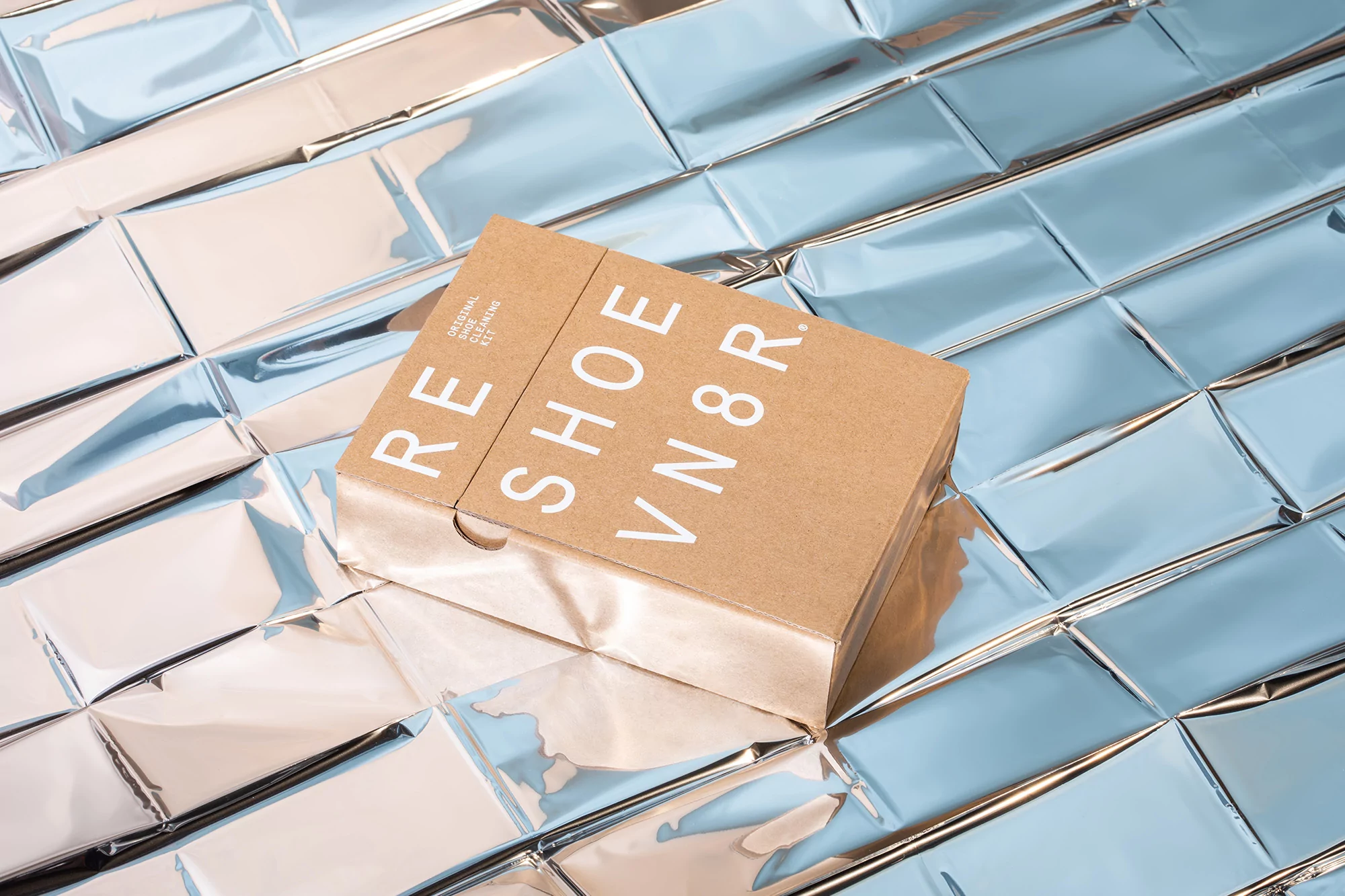 Angled shot of a RESHOEVN8R shoe cleaning kit box on a blue and silver metallic tile surface in a product studio shot.