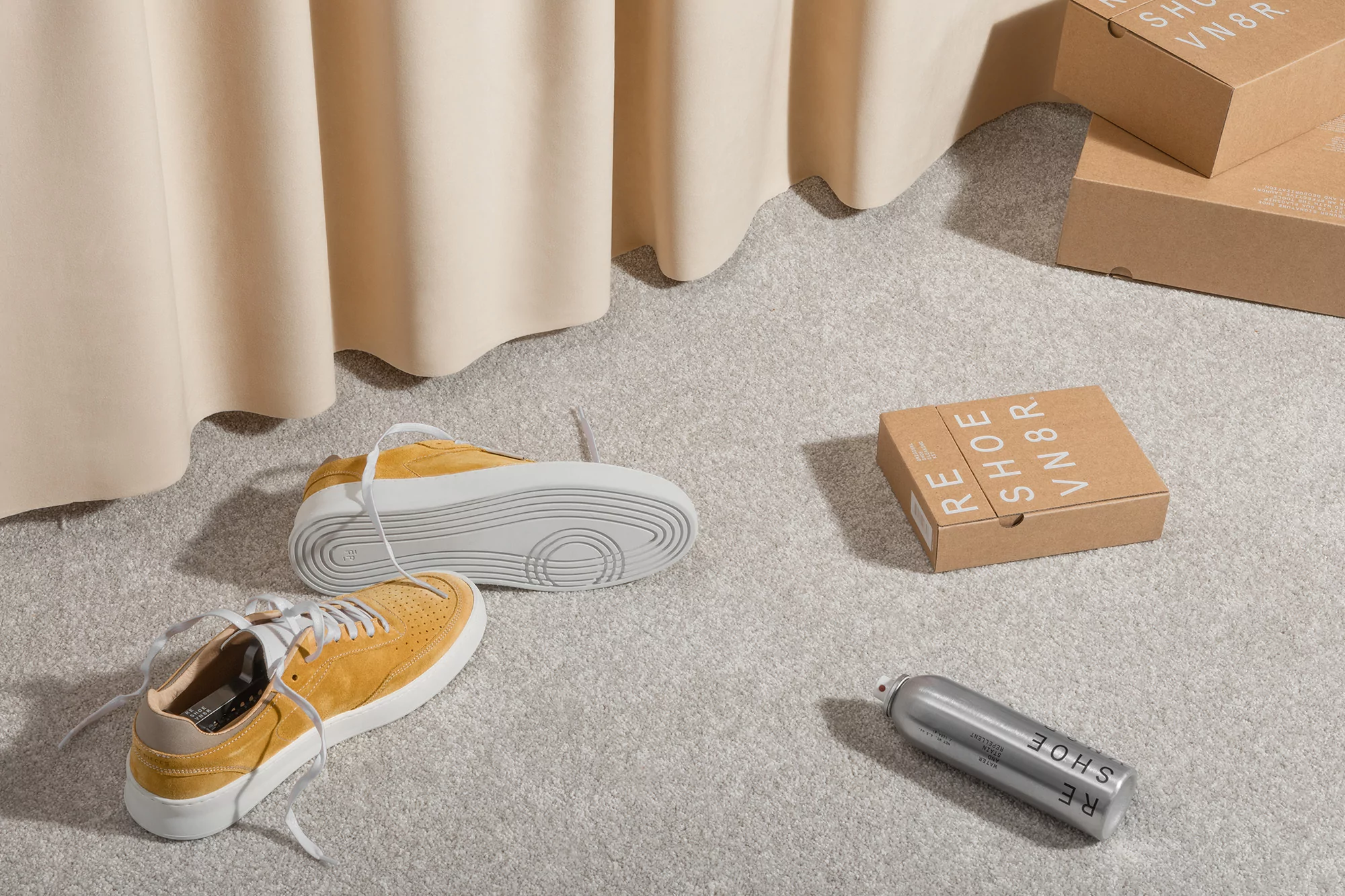 Elevated studio shot of mustard-colored sneakers, shoe cleaner, and boxes on a textured grey surface, complemented by draped cream fabric.
