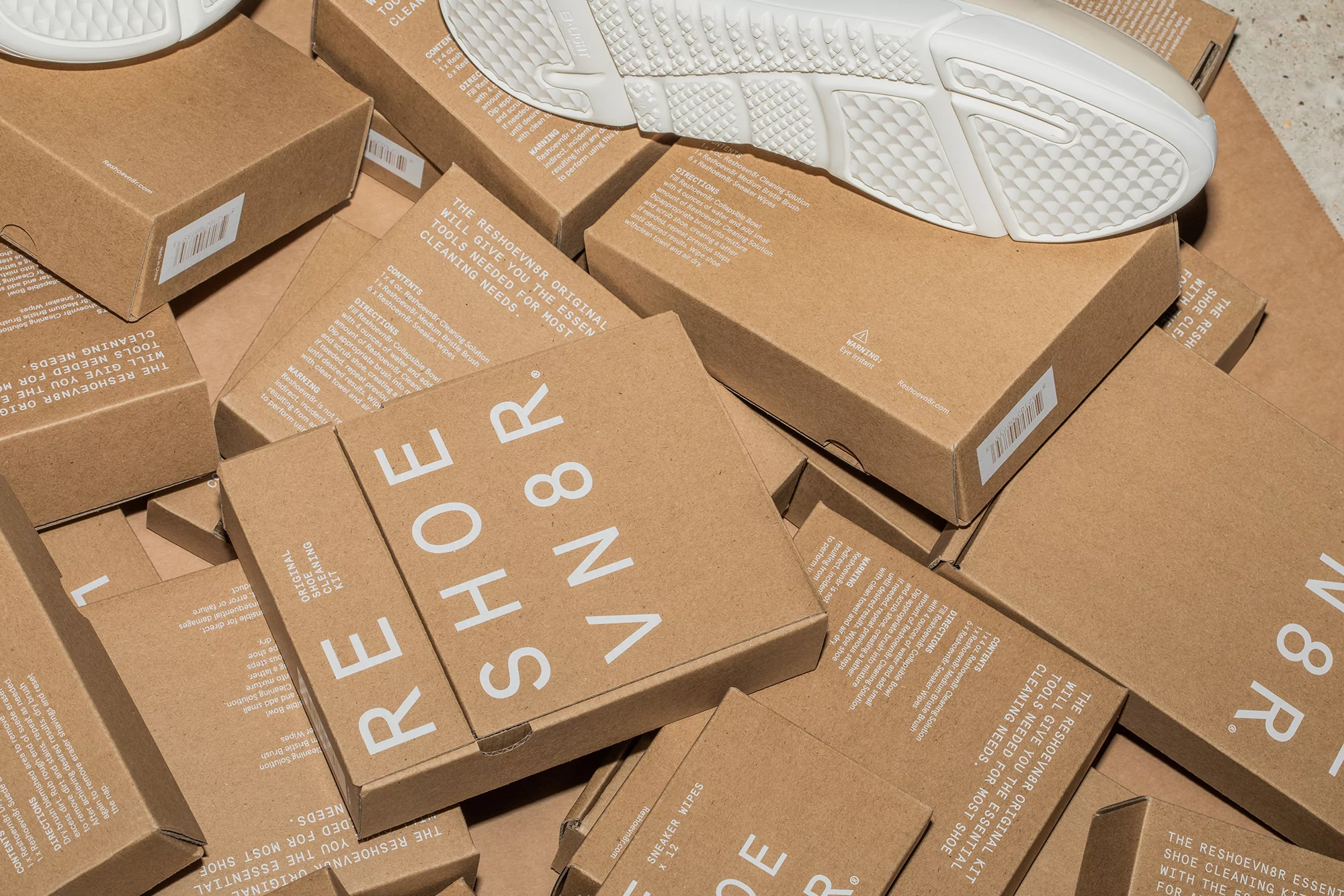 Pile of tan Reshoevn8r brand shoe cleaning kit boxes, with one white sneaker resting on top, and visible product text.