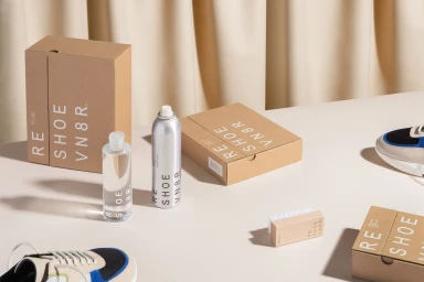 Reshoevn8r shoe care products, cardboard boxes, and sneakers arranged on a light surface against drapes