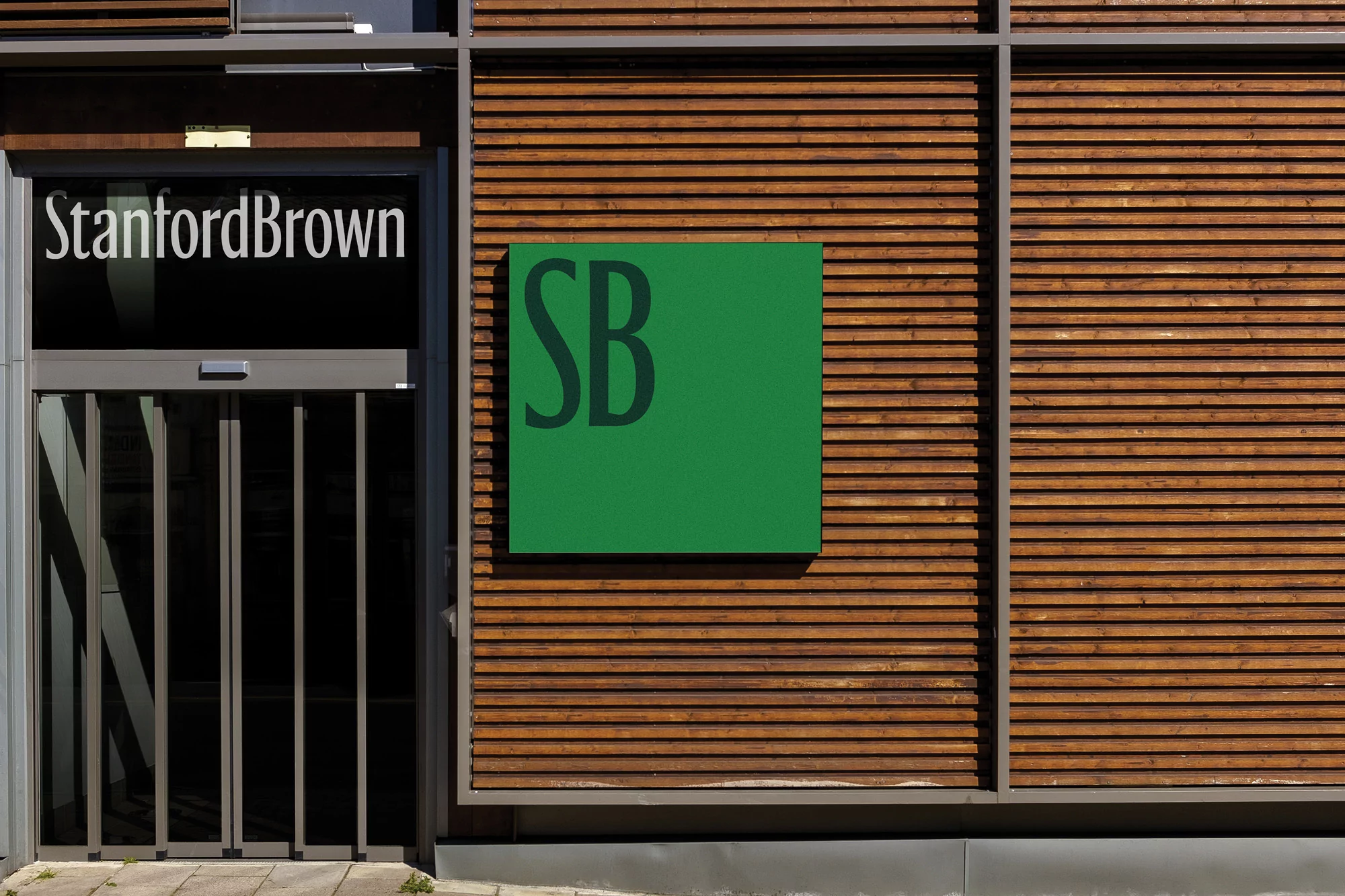 Exterior of commercial building featuring natural wood cladding, glass door with 'Stanford Brown' signage, and a green square logo.
