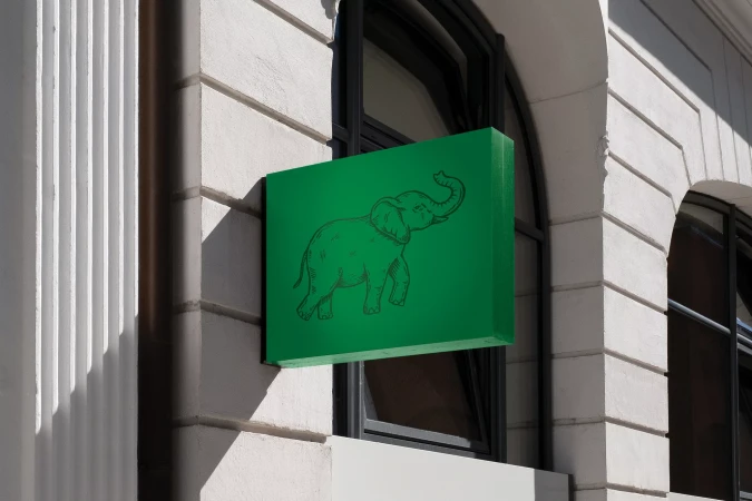 Green rectangular storefront sign featuring a black hand-drawn elephant graphic mounted on a white stone building exterior.