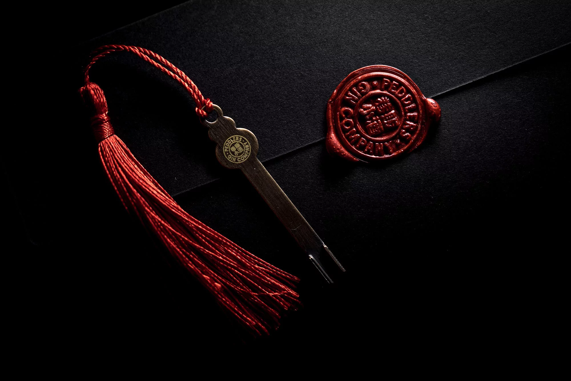 Still life image showcases a metal key with a red tassel, a wax seal, and a textured black surface, all under moody lighting.