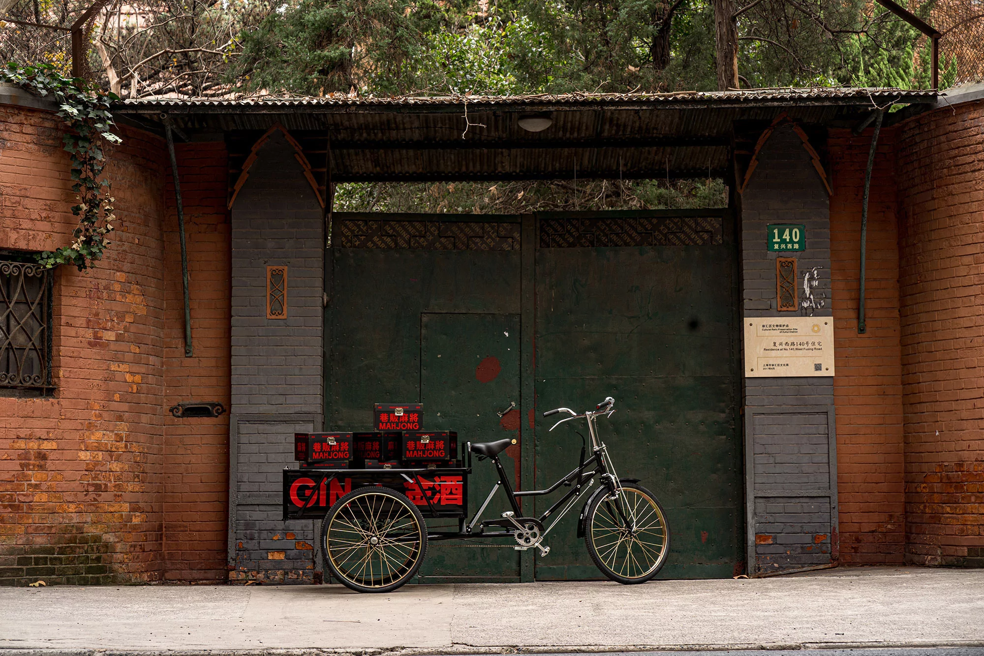 A vintage-style bicycle rickshaw parked against a brick building and dark green gate, with boxes marked 'Mahjong' loaded on the back.