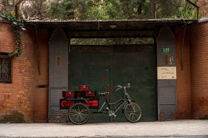 Black cargo tricycle with branded delivery boxes parked in front of a large green metal gate and weathered red brick wall