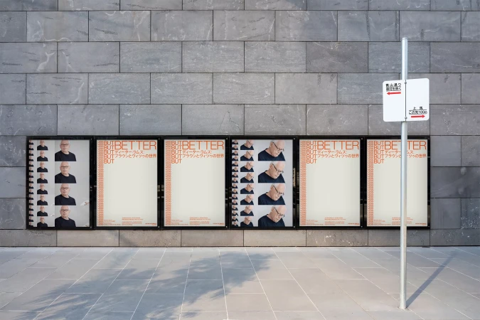 Outdoor view of several identical advertising posters on a grey brick wall with Japanese text next to a sign on a metal pole.