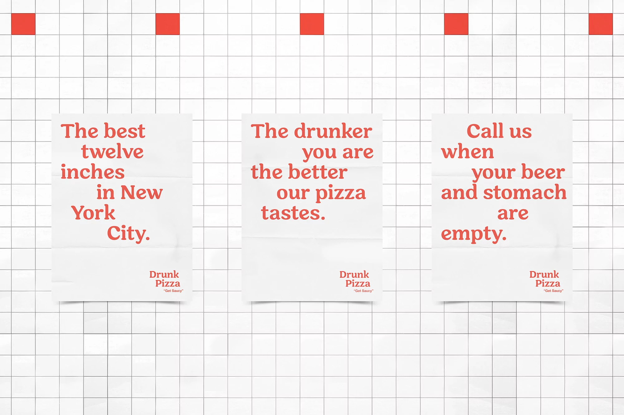 Three white posters with pizza advertisements hanging on a white grid tile wall with red squares, promoting Drunk Pizza in New York City.