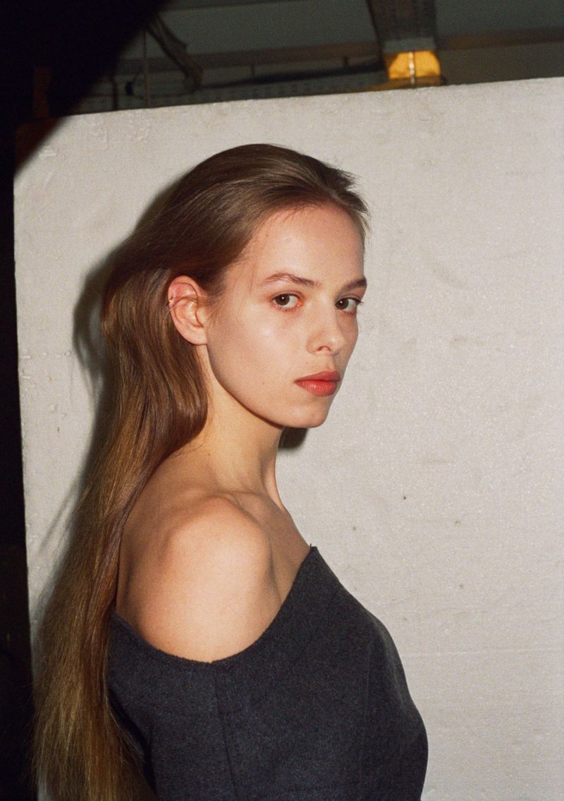 Portrait of a woman with long brown hair and fair skin wearing a dark grey dress, posing indoors with a neutral expression and soft lighting.