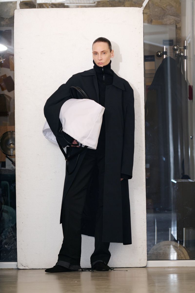 A slender adult fashion model in a black coat and pants holds a white bag indoors against a pale backdrop in a full-length shot.