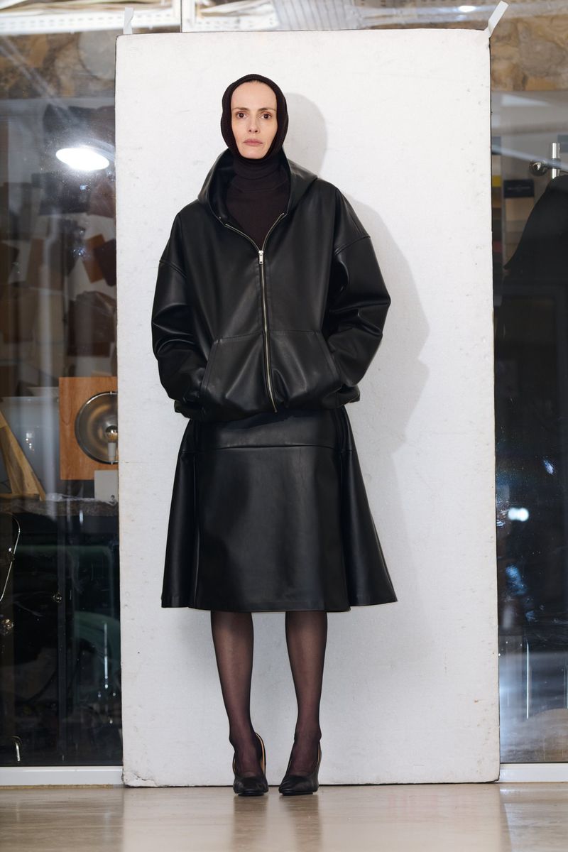 A woman poses in a black leather hooded jacket and skirt against a white backdrop, showcasing a minimalist fashion aesthetic.