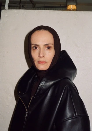 Portrait of an adult woman wearing a dark knitted head covering and a black faux leather hooded jacket in a brightly lit indoor space.