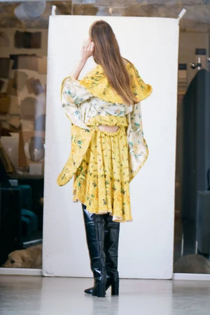 Model with long brown hair wearing a yellow floral dress and black thigh-high boots against a white backdrop.