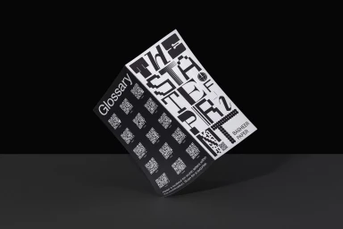 A rectangular printed brochure with bold graphic typography and QR codes tilted against a dark grey background.
