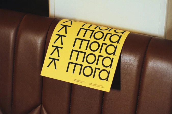 A yellow advertisement card with the word 'mora' repeatedly printed on it, placed on a brown, ribbed leather surface.