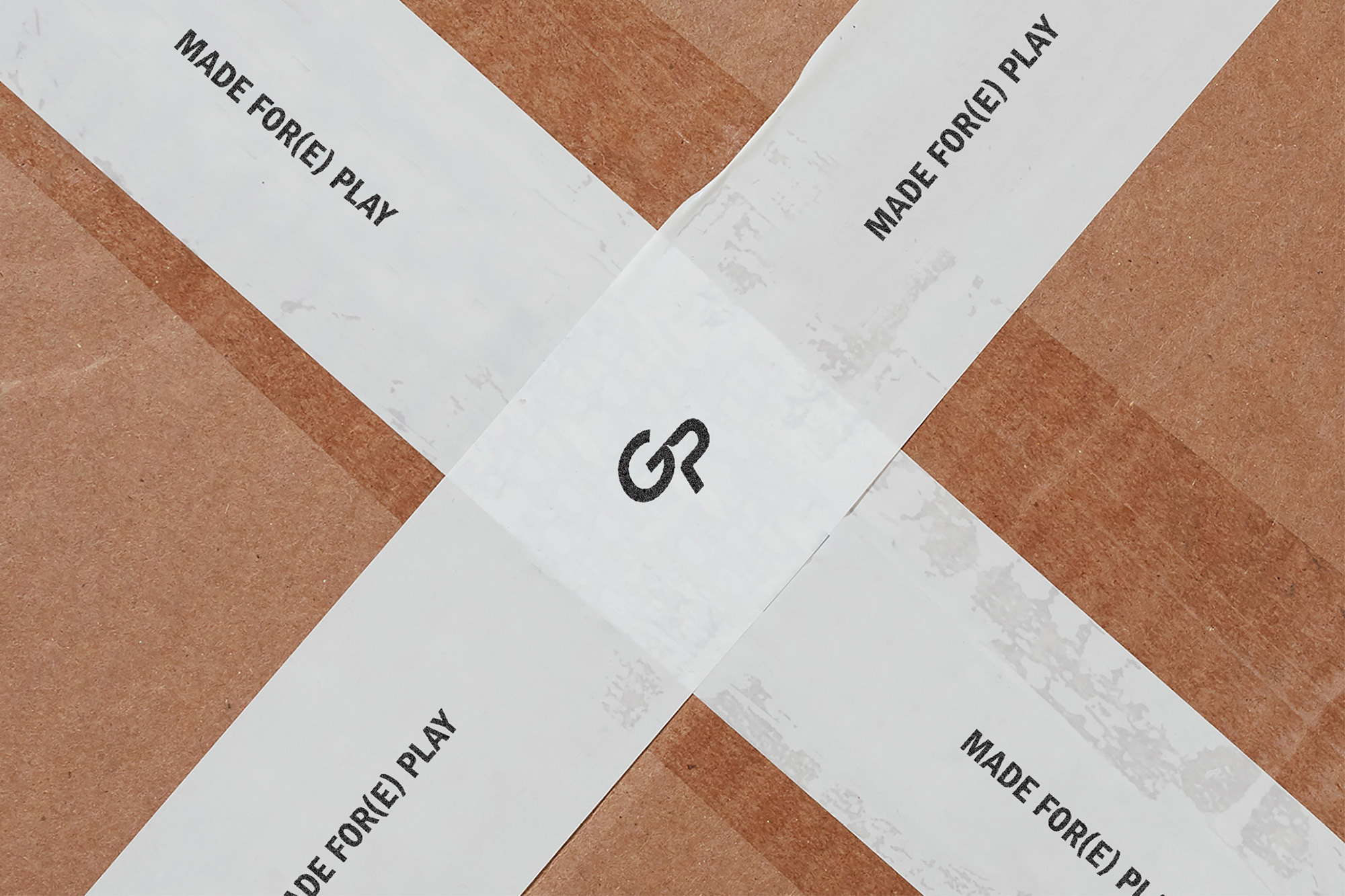 Close-up of a brown cardboard box sealed with white tape forming an X, with black text 'MADE FOR(E) PLAY' and a logo visible.