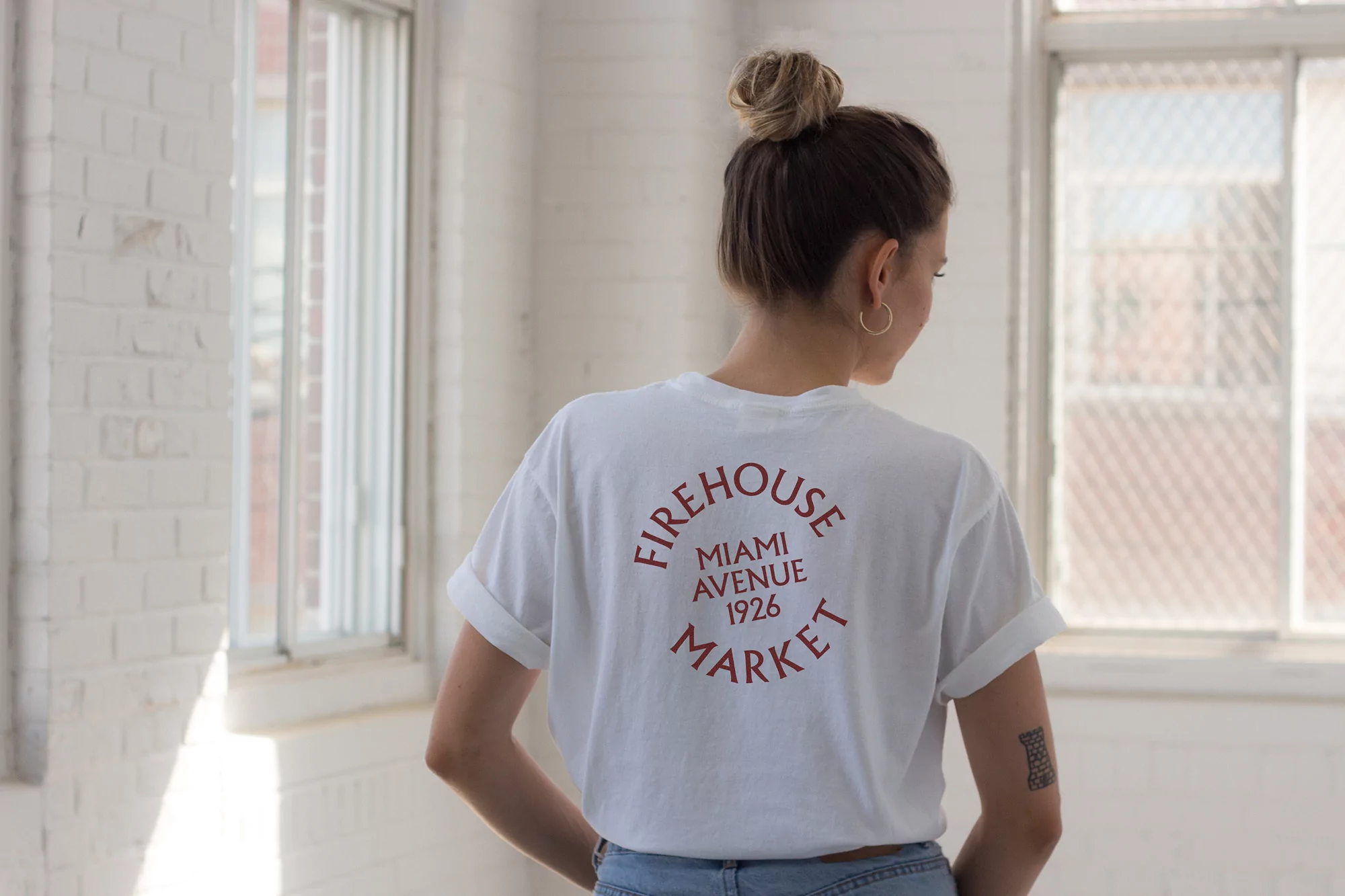 Back view of a slender woman with a topknot hairstyle and a white T-shirt standing in a light-filled interior by a window.