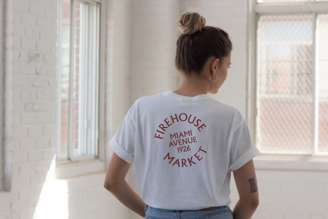 Back view of a slender woman with a topknot hairstyle and a white T-shirt standing in a light-filled interior by a window.