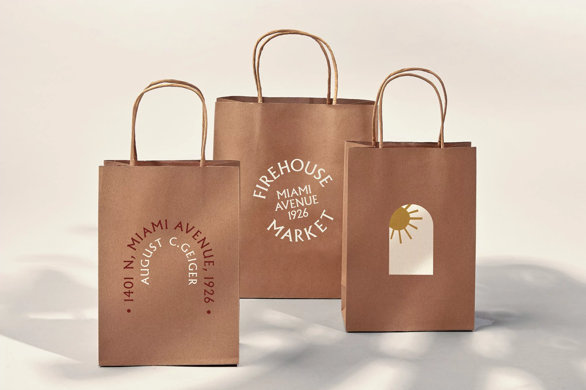 Three paper shopping bags with logo designs for Firehouse Miami Avenue Market and 1401 N August C. Geiger stand on a white surface.