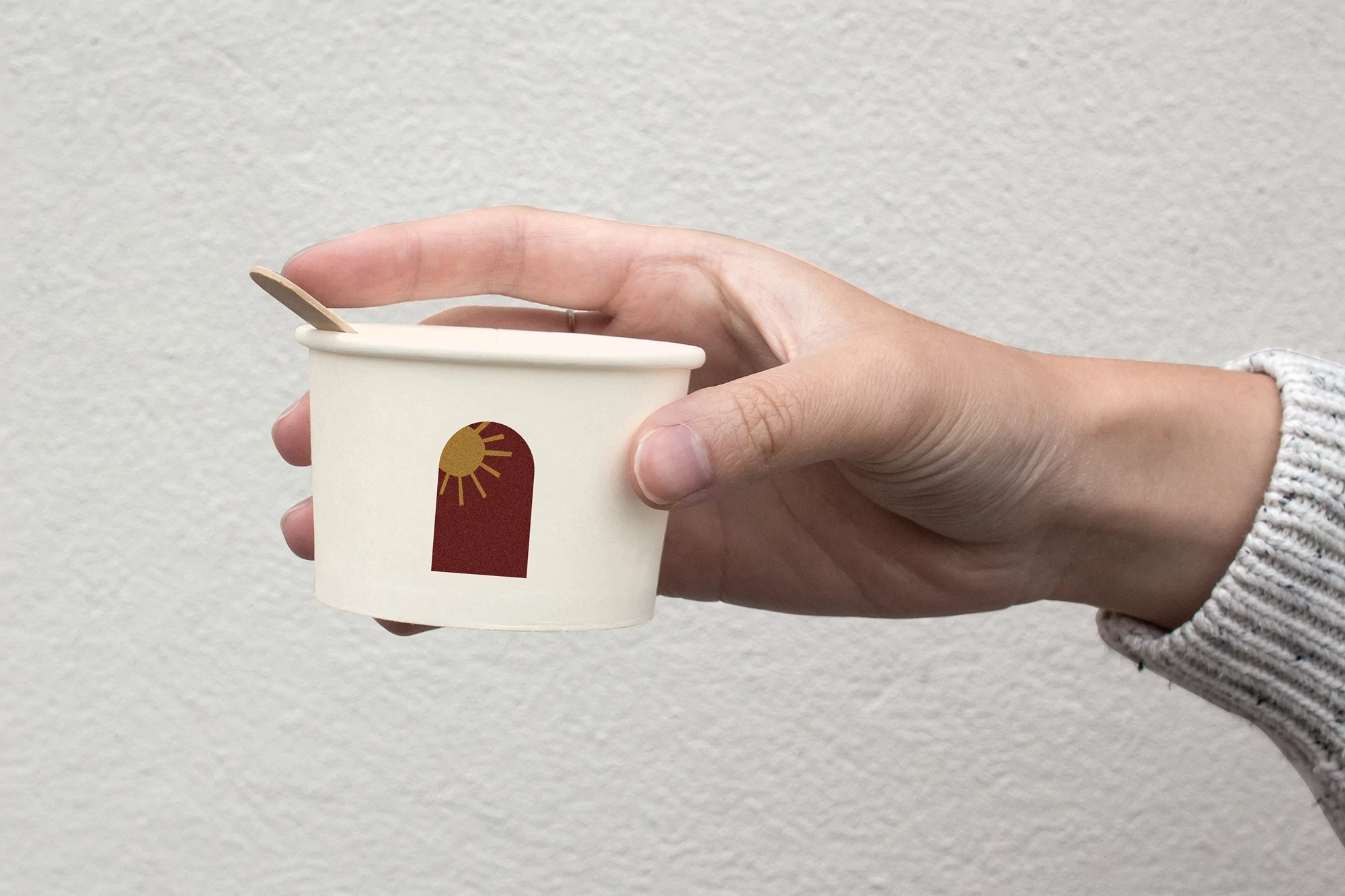 A hand holds a small, off-white paper cup with a wooden spoon inserted. The cup features a graphic with an arch design.