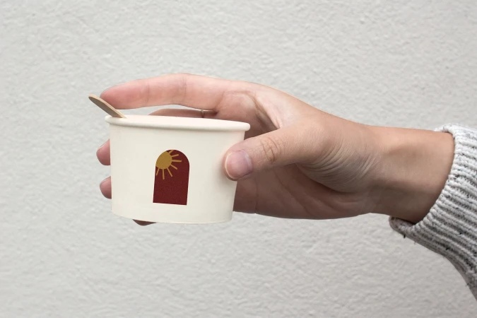 A hand holds a small, off-white paper cup with a wooden spoon inserted. The cup features a graphic with an arch design.