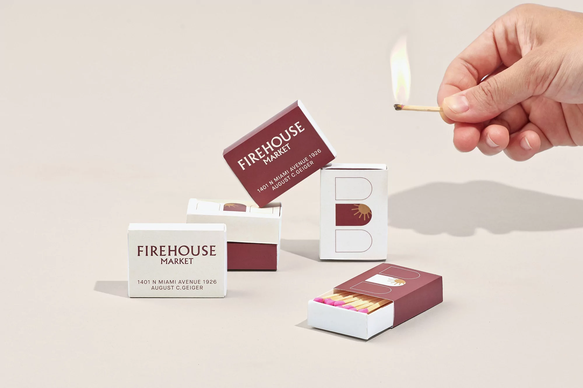 Stack of matchboxes with branding for Firehouse Market, one match lit and held in hand against a pale background.