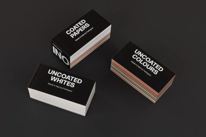 Close-up of three stacks of black paper sample books on a dark surface, featuring 'Coated Papers', 'Uncoated Whites' and 'Uncoated Colors'.