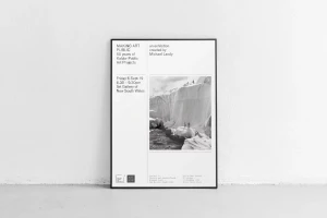 Black and white framed exhibition poster leaning against a plain white wall on a concrete floor.
