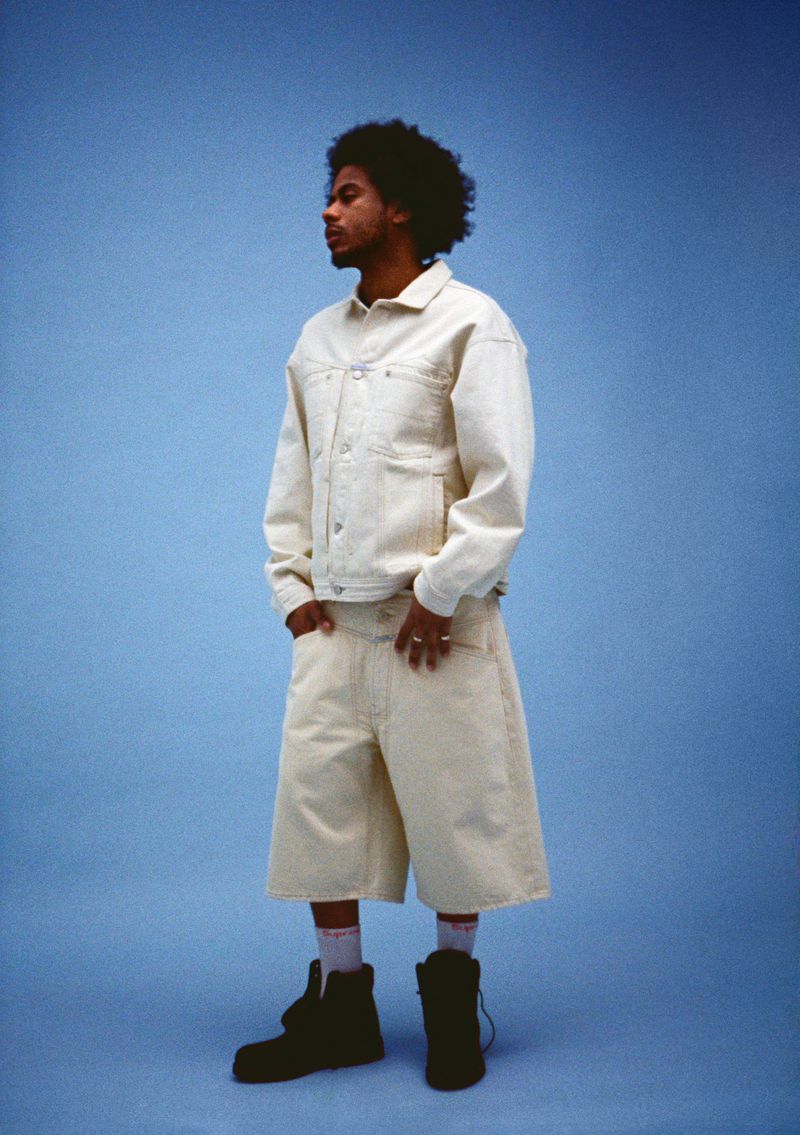 Adult male stands against a blue backdrop wearing an off-white denim jacket, shorts, and black boots for a fashion editorial shot.