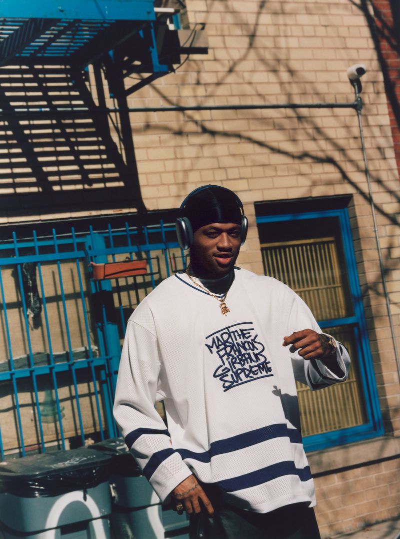 Young adult male in white graphic shirt with headphones in urban setting with brick wall and blue metal structure as background.